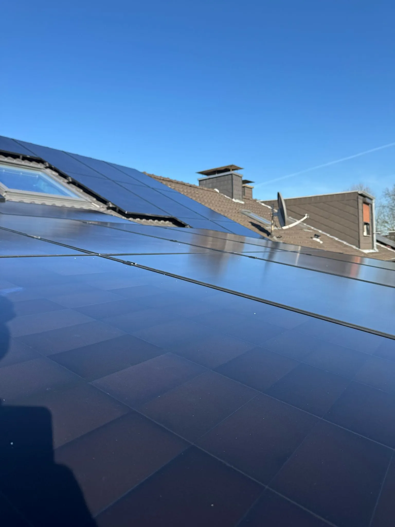 Vollflächig belegtes Ziegeldach mit schwarzen Photovoltaikmodulen, im Hintergrund Dachfenster und Schornstein